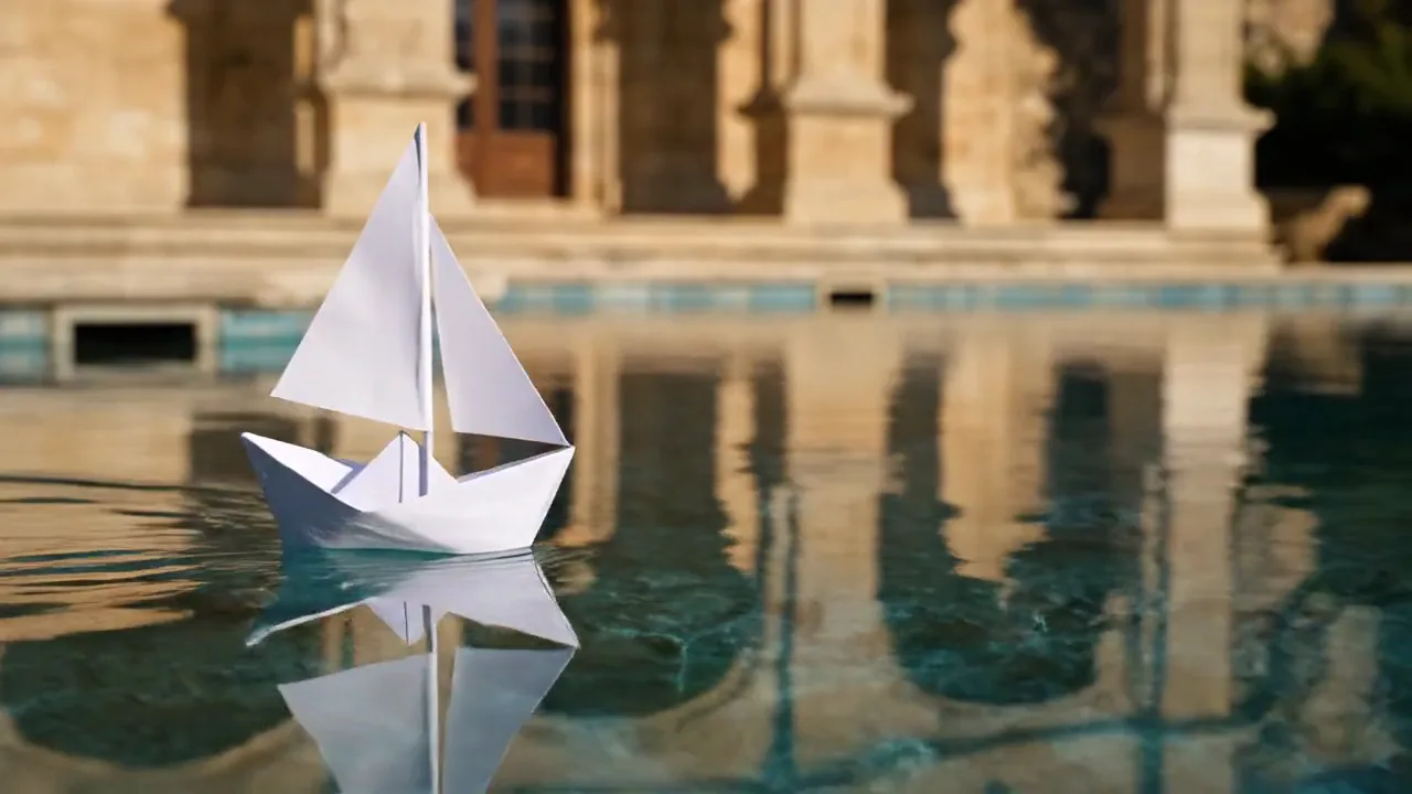 Still frame: A paper sailboat crossing a sunlit pool as the camera pushes in